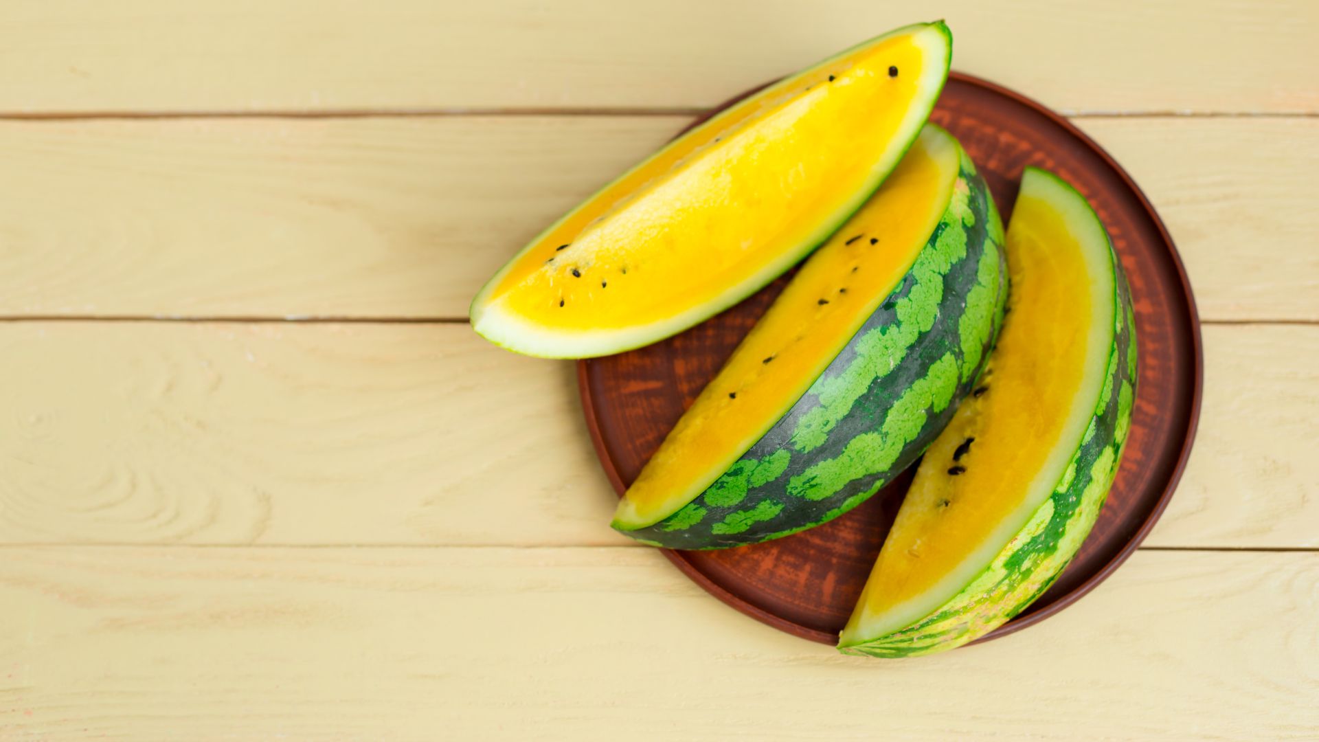 three freshly cut summer fruits slices of watermelon rest on a wooden circular thin plate on top of a manilla wooden table